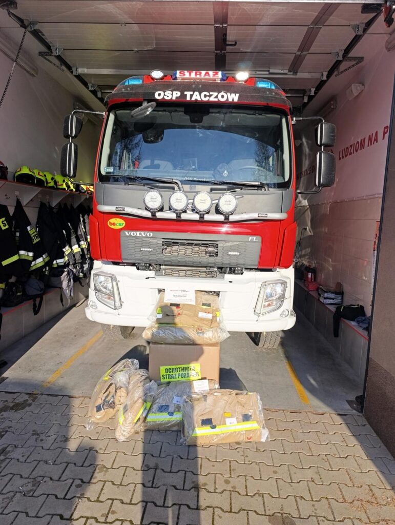 Samochód ratowniczo-gaśniczy OSP Taczów stojący w garażu, przed nim ułożone nowe ubrania specjalne strażackie otrzymane w ramach dofinansowania.