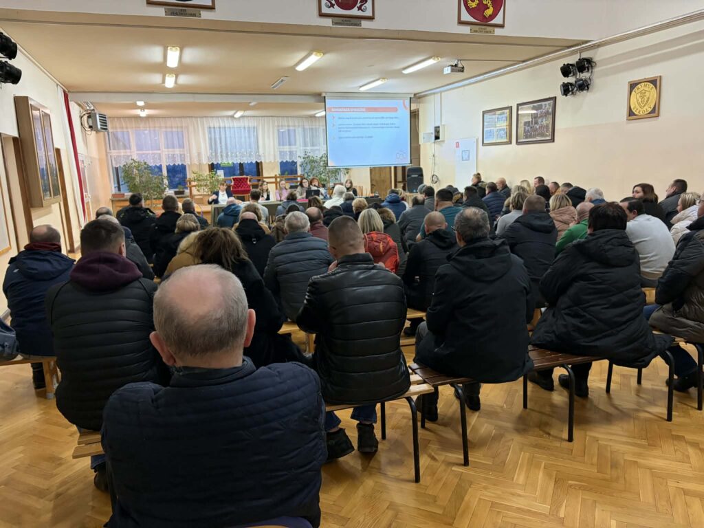 Uczestnicy konsultacji społecznych siedzą na ławkach w dużej sali. Na froncie widać panel prelegentów oraz ekran z prezentacją, a publiczność słucha wystąpienia