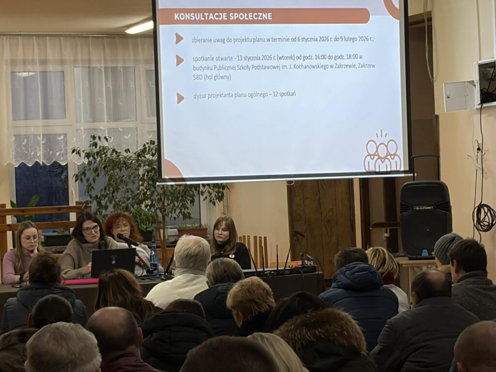 Panel prowadzących konsultacje społeczne siedzi przy stole przed publicznością. Za nimi wyświetlany jest slajd z informacjami o terminach konsultacji społecznych