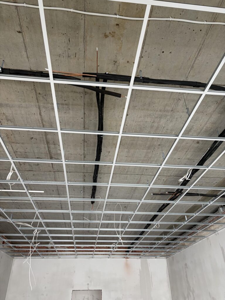 Konstrukcja sufitu podwieszanego z metalowych profili pod betonowym stropem, widoczne przewody elektryczne i rury instalacyjne przed zabudową