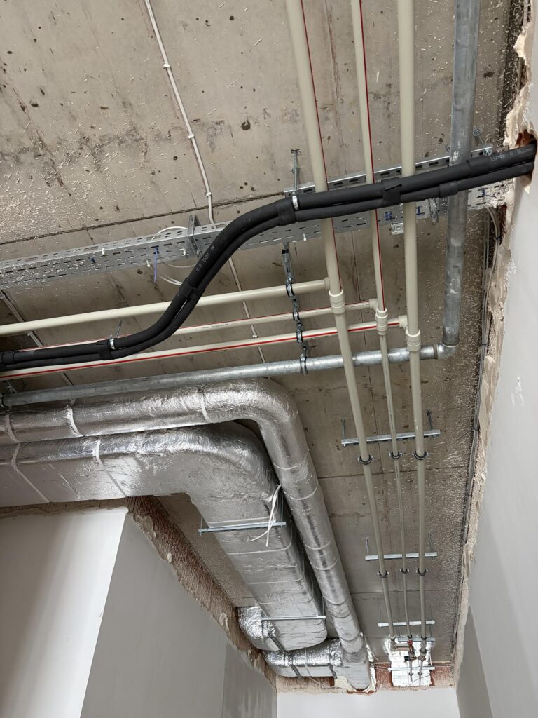 Odsłonięty betonowy strop z widocznymi instalacjami: rury hydrauliczne, kanały wentylacyjne w izolacji oraz koryta kablowe prowadzone wzdłuż sufitu
