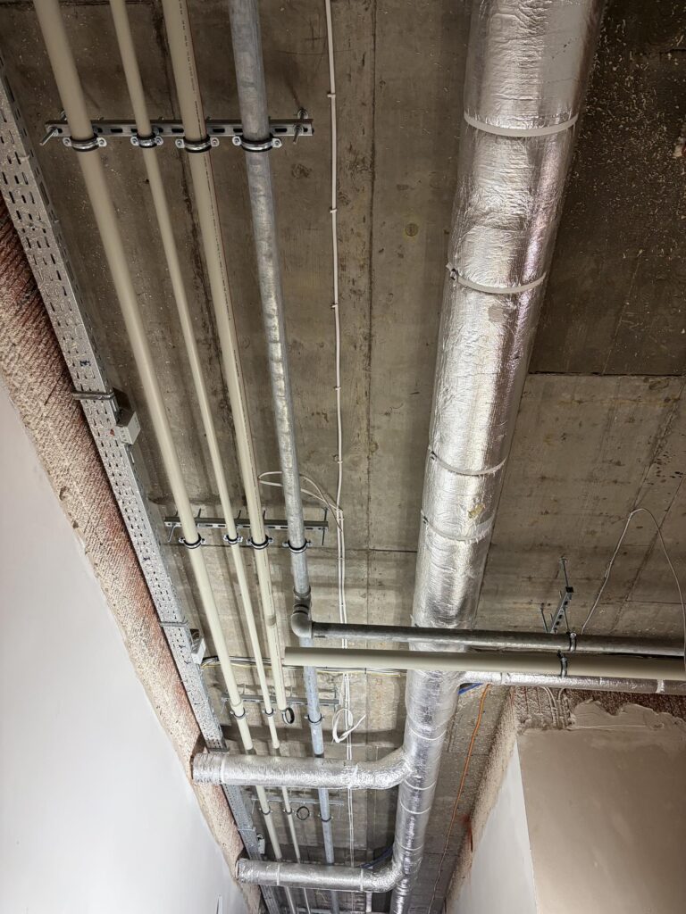 Odsłonięty betonowy strop z widocznymi instalacjami: rury hydrauliczne, kanały wentylacyjne w izolacji oraz koryta kablowe prowadzone wzdłuż sufitu