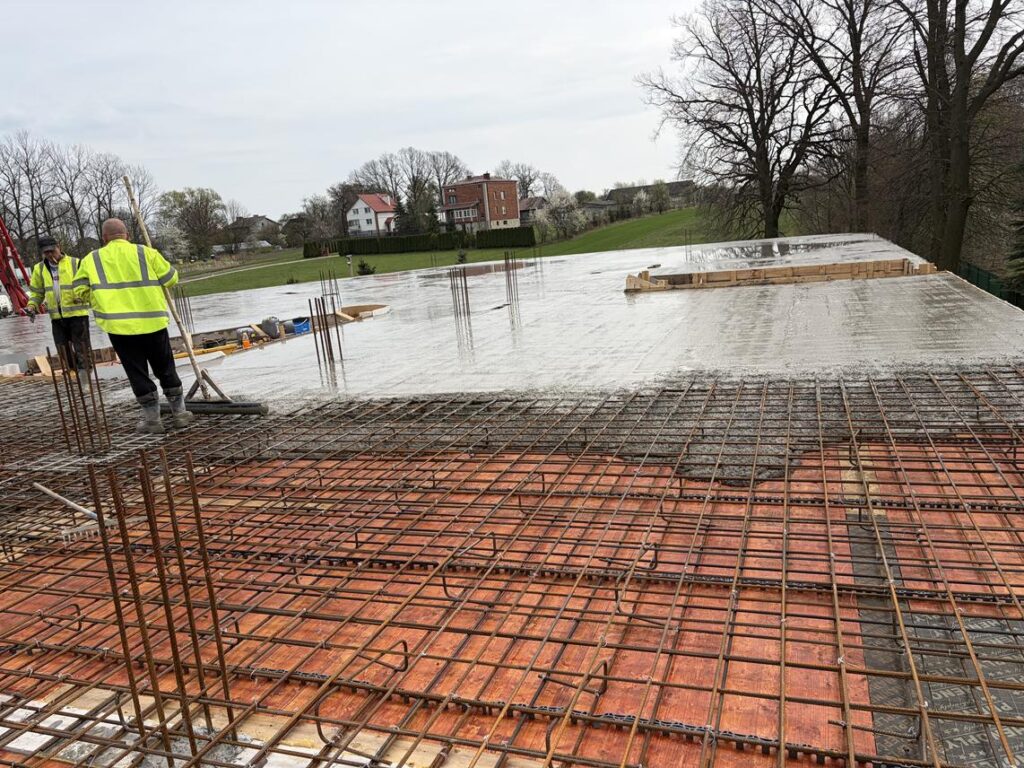 Na zdjęciu widać plac budowy, gdzie trwa wylewanie betonowego stropu. Na pierwszym planie znajduje się gęsta siatka zbrojeniowa z prętów stalowych ułożonych krzyżowo. W tle dwóch pracowników w odblaskowych kurtkach wyrównuje świeżo wylany beton na dużej, płaskiej powierzchni.