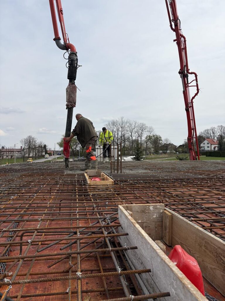 Zbrojony strop na budowie – dwóch pracowników wlewa beton przy użyciu pompy. Widoczna gęsta siatka prętów stalowych oraz elementy szalunku.