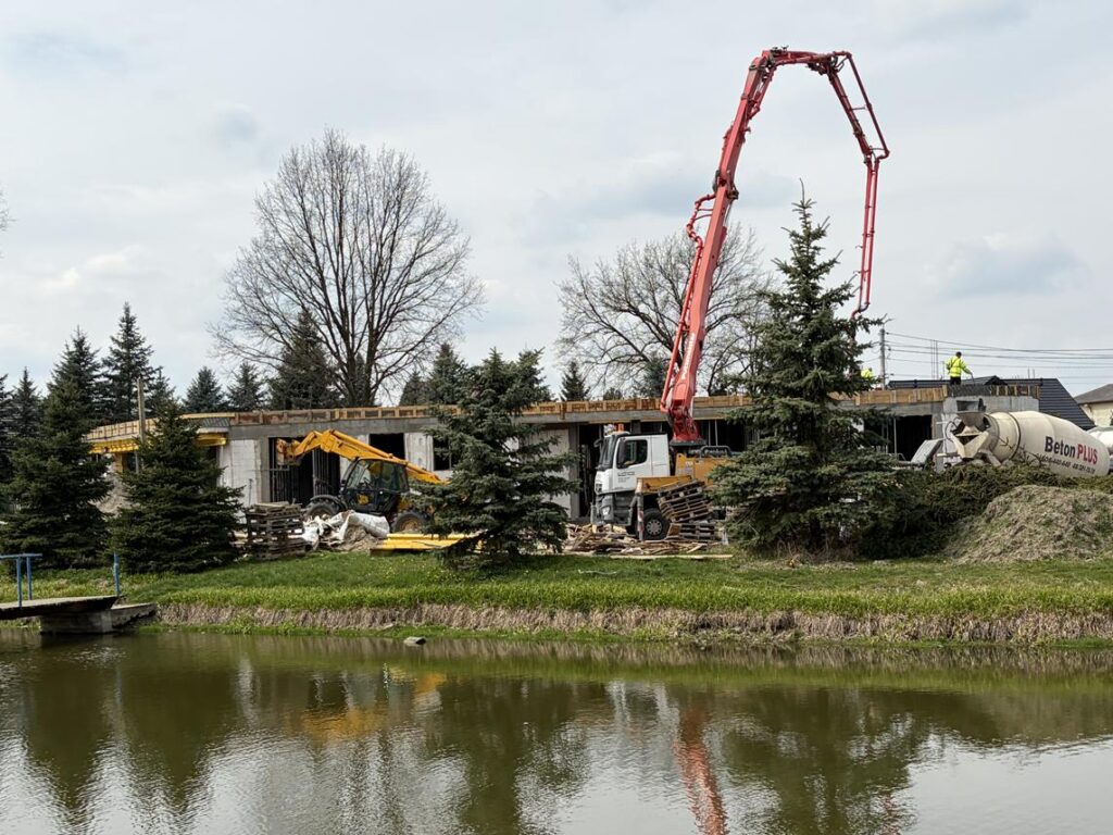 Plac budowy nad wodą – betonowy budynek w trakcie wznoszenia. Widoczna pompa do betonu z długim czerwonym ramieniem oraz betoniarka. Wokół znajdują się drzewa i trawa.