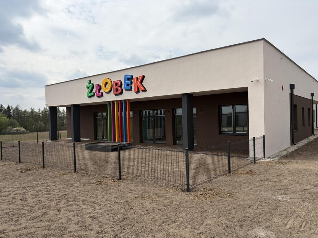 Wejście do nowoczesnego budynku żłobka z napisem „ŻŁOBEK” w kolorowych literach nad zadaszeniem. Przed budynkiem znajduje się ogrodzony teren z nieutwardzoną nawierzchnią.