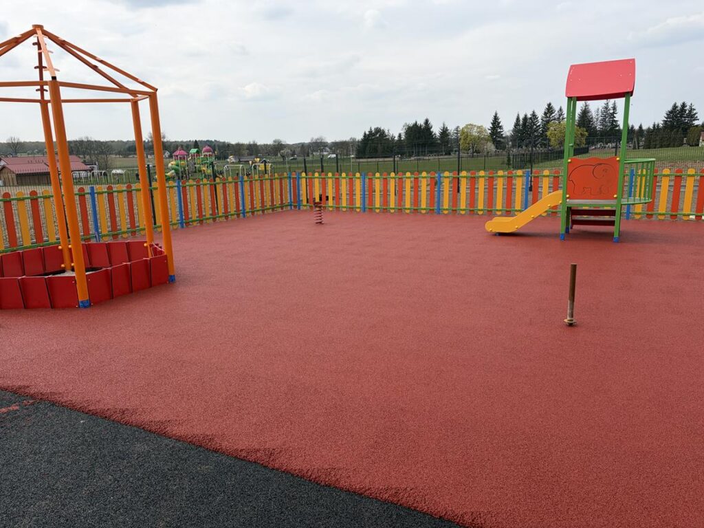 Fragment placu zabaw z bezpieczną, gumową nawierzchnią w kolorze czerwonym. Na środku znajduje się sprężynowy bujak, a z boku mała zjeżdżalnia z kolorową konstrukcją. Całość ogrodzona jest barwnym płotkiem, a w tle widać drzewa i otwartą przestrzeń.