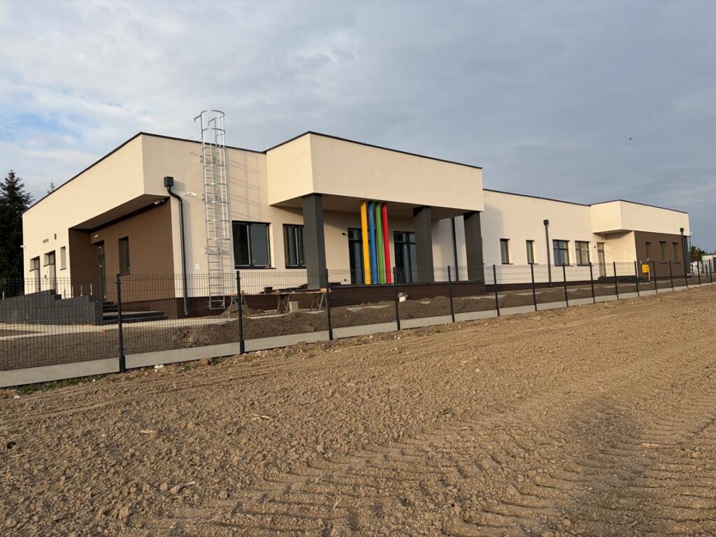 Nowoczesny, parterowy budynek żłobka o jasnej elewacji, z płaskim dachem i kolorowymi pionowymi elementami dekoracyjnymi przy wejściu. 