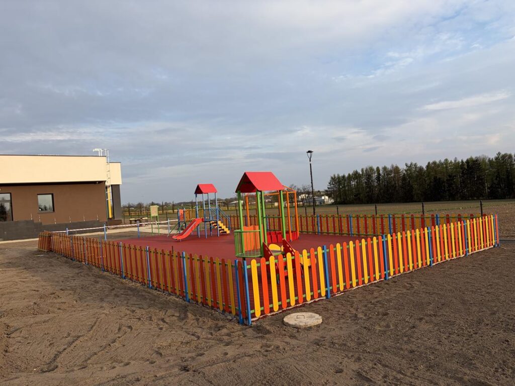 Plac zabaw otoczony kolorowym ogrodzeniem z czerwono-pomarańczową nawierzchnią. Na terenie znajdują się małe zjeżdżalnie, konstrukcje do wspinania oraz sprężynowy bujak. W tle widać nowoczesny, jasny budynek z płaskim dachem oraz otwarte tereny zielone i drzewa.