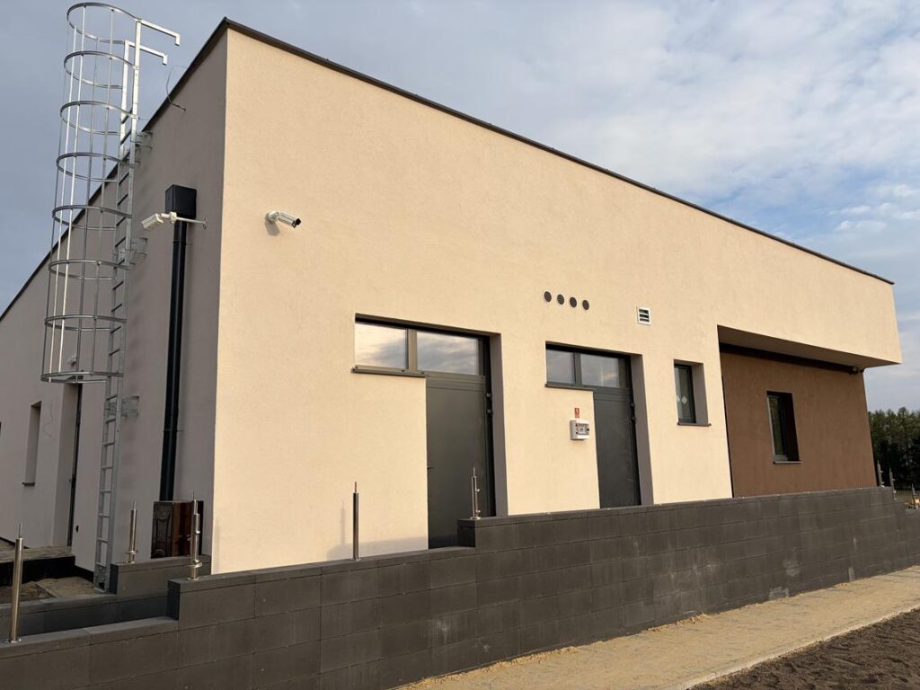 Nowoczesny, parterowy budynek o jasnej elewacji i płaskim dachu. Widoczne są drzwi wejściowe, okna, monitoring oraz metalowa drabina prowadząca na dach. Budynek otoczony jest utwardzonym terenem i niskim murkiem.