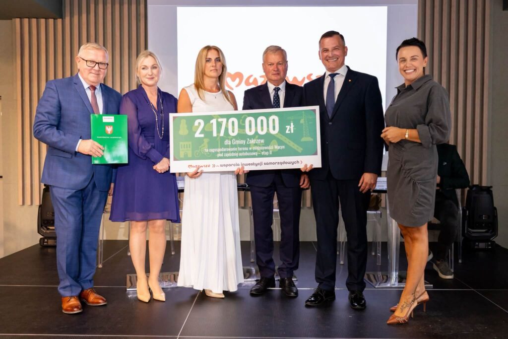Sześć osób stoi na scenie podczas uroczystości wręczenia dofinansowania. Trzy osoby w środku trzymają dużą tablicę z informacją o przyznaniu 2 170 000 zł dla Gminy Zakrzew na zagospodarowanie terenu w miejscowości Wacyn pod zajezdnię autobusową (etap II). Po lewej stronie mężczyzna w niebieskim garniturze trzyma zieloną teczkę z herbem województwa. W tle widoczny jest ekran z logo programu „Mazowsze” oraz stół konferencyjny.