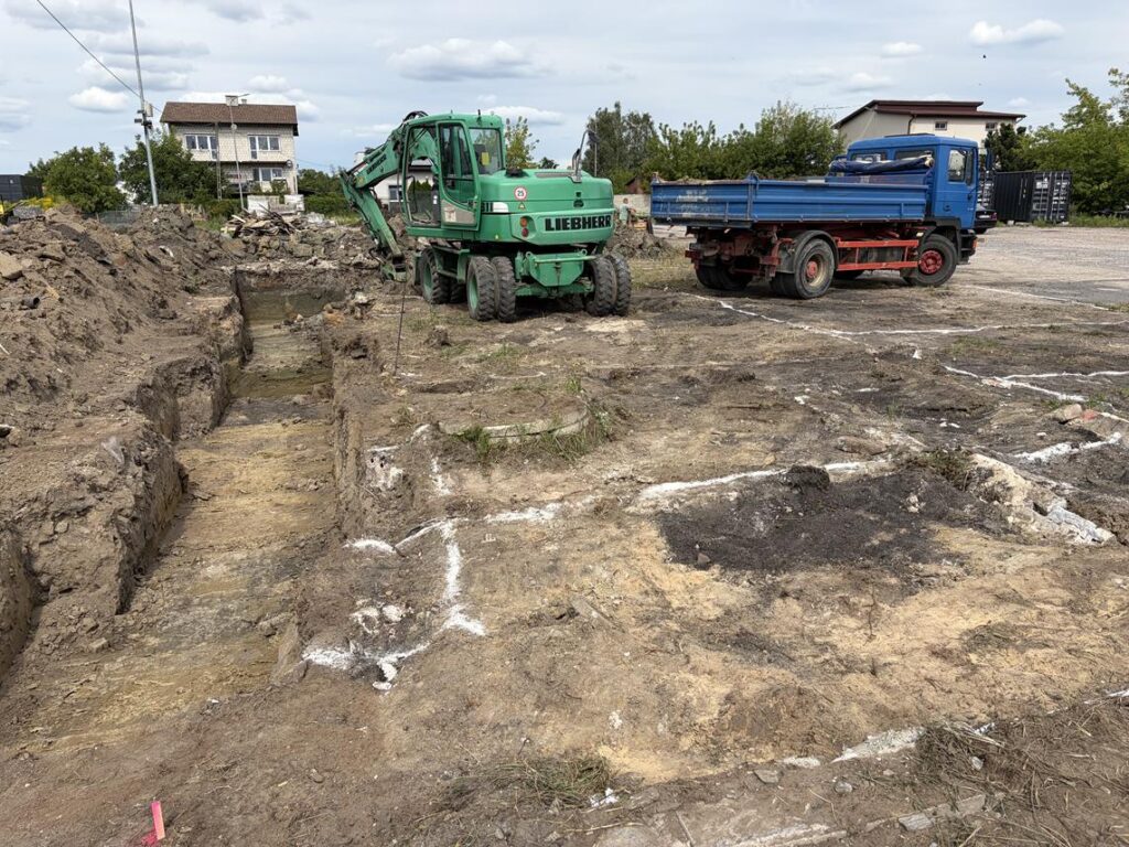 Koparka Liebherr i niebieska wywrotka pracują na terenie robót ziemnych; widoczne są głębokie wykopy, pryzmy ziemi oraz oznaczenia geodezyjne na gruncie.