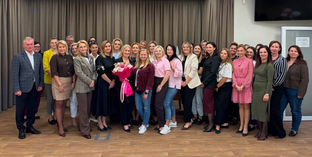 Na zdjęciu widać pracowników Urzędu Gminy w Zakrzewie - około 25–30 osób, ustawionych wspólnie do pamiątkowej fotografii z okazji pożegnania pracownika. W centrum stoi kobieta trzymająca duży bukiet kwiatów w różowej oprawie. Większość osób to kobiety, a w grupie jest także kilku mężczyzn. Uczestnicy są elegancko i półformalnie ubrani — widać m.in. marynarki, sukienki, spódnice, koszule i swetry. Wszyscy stoją przed dużymi, zasłoniętymi zasłonami. W tle po prawej stronie znajduje się ekran oraz drzwi z informacją umieszczoną na ścianie. Atmosfera zdjęcia jest uroczysta, ale jednocześnie swobodna i radosna — wiele osób się uśmiecha i pozuje blisko siebie. Zdjęcie wykonano w sali konferencyjnej. 