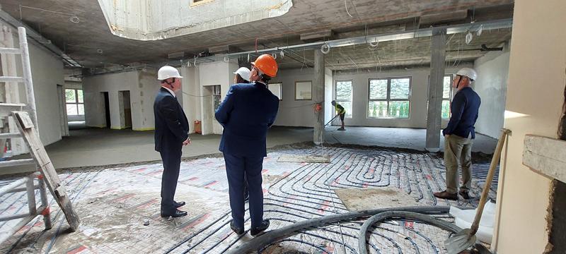 Na zdjęciu widać wnętrze budowanego budynku. Trzy osoby w kaskach ochronnych oglądają postęp prac.
Widoczne są m.in.:
rozłożone na podłodze pętle rur ogrzewania podłogowego, przygotowane przed wykonaniem wylewki, odsłonięte instalacje elektryczne i przewody prowadzone pod sufitem, surowy betonowy strop i słupy konstrukcyjne, niewykończone ściany i pomieszczenia, drabina oraz narzędzia budowlane, w głębi pracownik wykonujący prace przy podłodze.
Dwie osoby są ubrane w garnitury, a trzecia w odzież roboczą. Wygląda to na oględziny placu budowy podczas wizyty osób nadzorujących lub związanych z inwestycją.