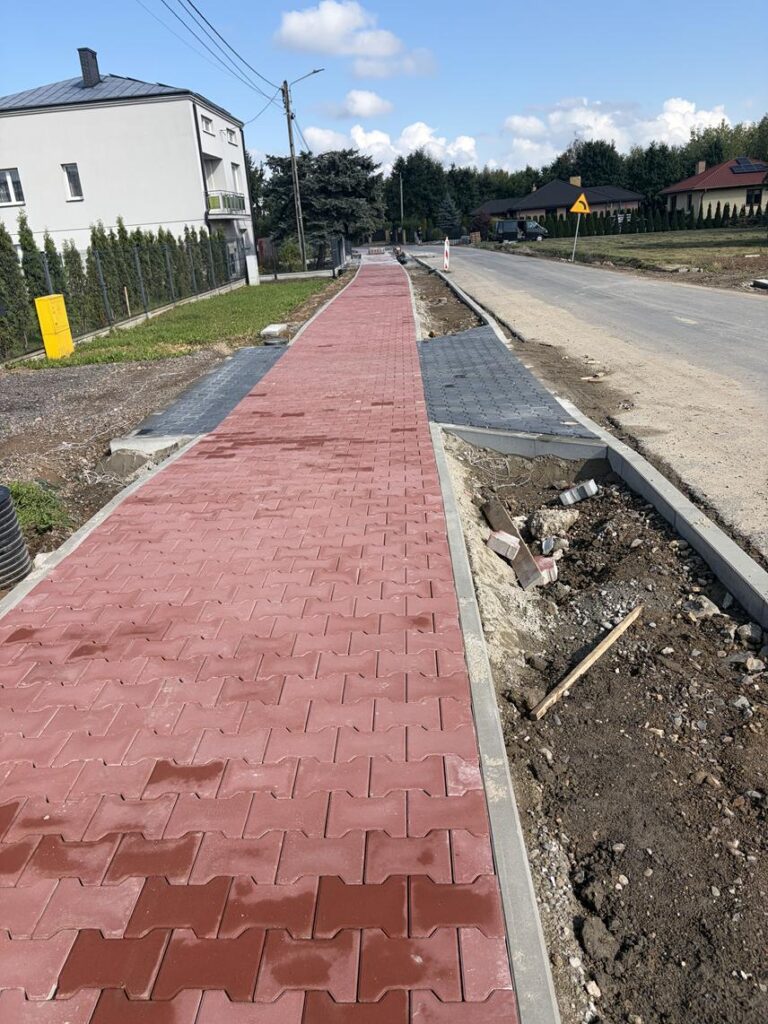 Nowo ułożony czerwony chodnik z kostki brukowej wzdłuż remontowanej drogi osiedlowej; po bokach widoczne szare pasy z kostki, niezagospodarowane pobocze, domy jednorodzinne, drzewa i znak ostrzegawczy.