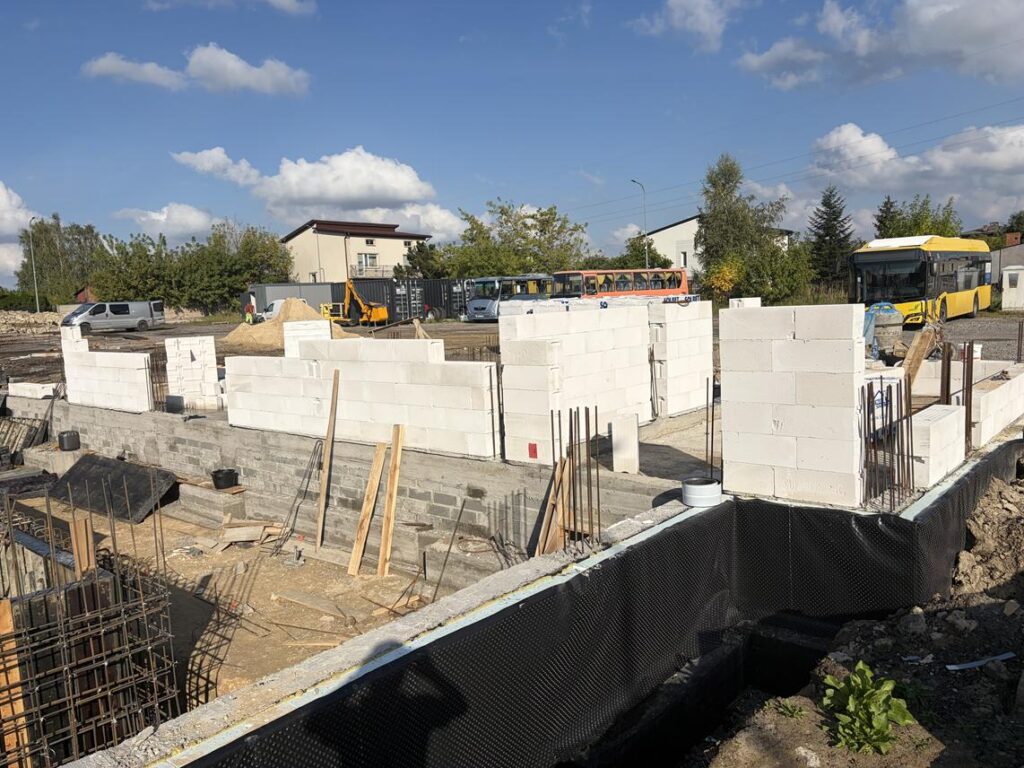 Plac budowy z wznoszonymi ścianami z białych bloczków, widocznym zbrojeniem fundamentów i wykopami. W tle znajdują się samochody, sprzęt budowlany, drzewa oraz żółty autobus