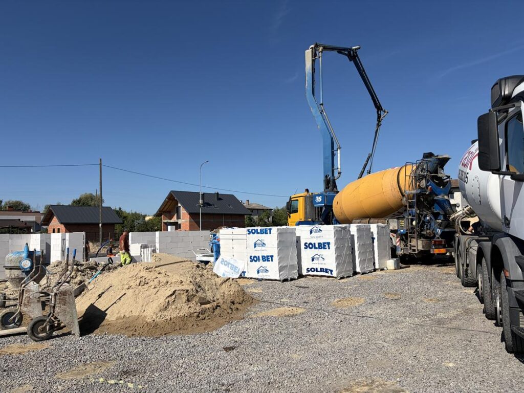 Plac budowy domu podczas prac fundamentowych. Na pierwszym planie znajdują się materiały budowlane, piasek i kruszywo, a w tle samochód z betonomieszarką i pompą do betonu oraz pracownicy budowy.