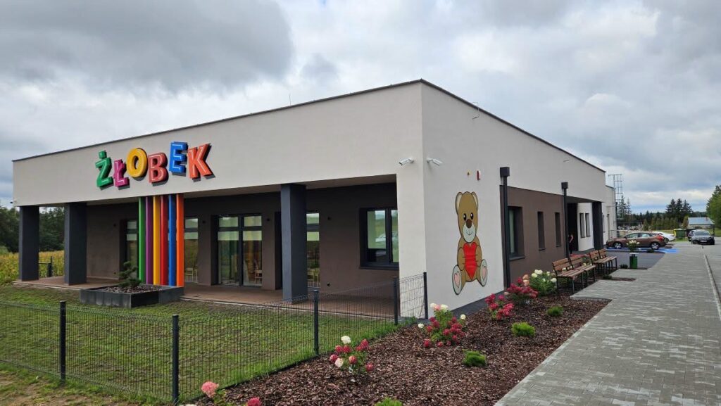 Na zdjęciu widać nowoczesny, parterowy budynek żłobka z dużym napisem „ŻŁOBEK” na elewacji. Budynek ma jasną, kremową fasadę z ciemnymi akcentami przy wejściach i duże okna. Przed wejściem znajdują się kolorowe pionowe elementy dekoracyjne, a na bocznej ścianie namalowano sympatycznego misia z czerwonym sercem. Teren wokół jest starannie zagospodarowany — widać ogrodzenie, trawnik, rabaty z kwitnącymi różami oraz ławki.
Po prawej stronie znajduje się wybrukowany chodnik i parking z kilkoma samochodami. W tle widać drzewa oraz zachmurzone niebo.