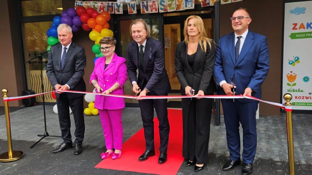 Na zdjęciu widać uroczyste otwarcie placówki żłobka. Przed wejściem stoi pięć osób, które trzymają nożyczki przy czerwono-białej wstędze, przygotowując się do jej przecięcia.
W centrum ustawiono czerwony dywan prowadzący do wejścia.
W tle znajduje się kolorowa dekoracja z balonów w kształcie łuku.
Nad drzwiami zawieszono baner z napisem wyglądającym na „… NAS…”, prawdopodobnie związanym z nazwą lub hasłem uroczystości.
Po prawej stronie widoczny jest baner z napisami m.in. „ZAK…” oraz „NASZ PRZ…”, a niżej grafiki kwiatów i motyli.
Uczestnicy są elegancko ubrani, a całość ma charakter oficjalnej uroczystości inaugurującej działalność placówki.
Widać również mikrofon i nagłośnienie, co sugeruje, że podczas wydarzenia odbywały się przemówienia.
Atmosfera zdjęcia jest uroczysta i jednocześnie radosna.