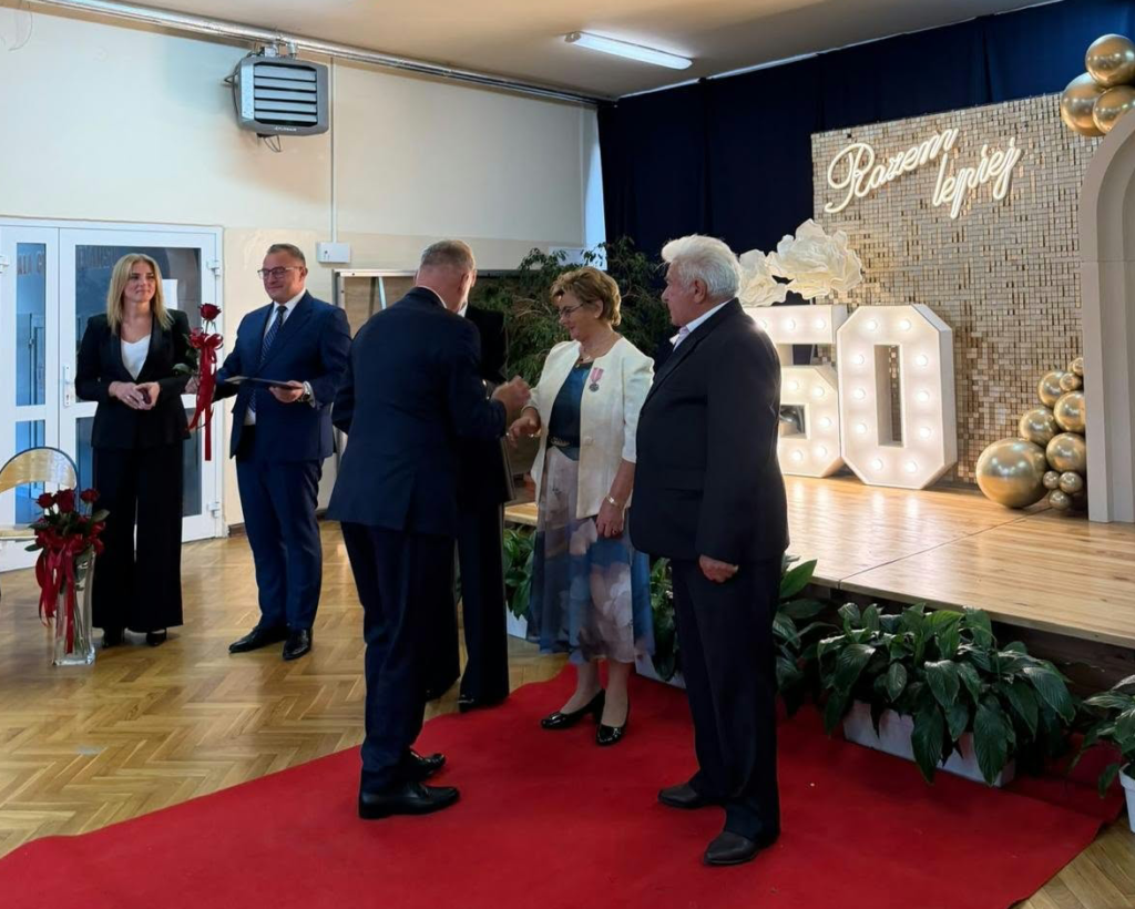 Podczas uroczystości jubileuszu długoletniego pożycia małżeńskiego przedstawiciel organizatorów wręcza jubilatce czerwoną różę. U jej boku stoi mąż, odznaczony medalem, z dumą i wzruszeniem uczestniczący w tej wyjątkowej chwili. W tle widoczny jest elegancki napis „Razem najlepiej”, który doskonale oddaje charakter spotkania.
To piękny gest uznania dla wspólnie przeżytych lat, wzajemnej miłości, wierności i przywiązania. Uroczystość przebiega w podniosłej, a zarazem serdecznej atmosferze, podkreślając, że prawdziwa miłość i trwałość małżeństwa zasługują na szczególne wyróżnienie.