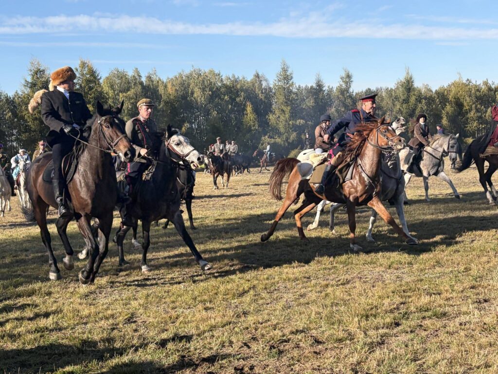 Na zdjęciu przedstawiono grupę jeźdźców poruszających się konno po rozległej, jesiennej łące. Uwagę przyciągają osoby ubrane w historyczne stroje, ciemne mundury, wysokie buty oraz charakterystyczne nakrycia głowy, co nadaje wydarzeniu klimat dawnej epoki. Na pierwszym planie widać kilka koni o różnym umaszczeniu — gniadym, siwym i kasztanowatym. Jeźdźcy pewnie trzymają wodze i przemieszczają się w różnych kierunkach, a w tle można dostrzec kolejnych uczestników konnego spotkania. Trawa jest miejscami wysuszona i przydeptana, a długie cienie wskazują na nisko padające promienie słońca. W oddali rozciąga się pas drzew o jesiennych barwach, nad którym góruje błękitne niebo.