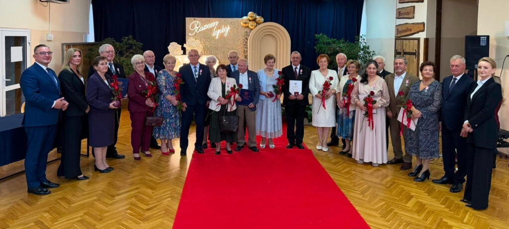 Na zdjęciu widzimy uroczyste spotkanie zorganizowane z okazji jubileuszu długoletniego pożycia małżeńskiego. Na środku sali, na tle granatowych zasłon, przygotowano elegancką dekorację z napisem „Razem najlepiej”, złotymi balonami oraz ozdobną ścianką.
Na czerwonym dywanie stoi grupa kilkudziesięciu osób – jubilatów, którzy otrzymali odznaczenia i pamiątkowe dyplomy, a także przedstawiciele władz lub organizatorzy uroczystości. Odświętny charakter wydarzenia podkreślają eleganckie stroje, czerwone róże wręczone uczestnikom oraz uśmiechy i serdeczna atmosfera.
To wyjątkowa chwila, będąca wyrazem uznania dla wieloletniej miłości, wzajemnego szacunku, wierności i wspólnie przeżytych lat. Zdjęcie upamiętnia piękny jubileusz oraz historie małżeństw, które przez lata budowały swoje rodziny i pielęgnowały łączące je więzi.