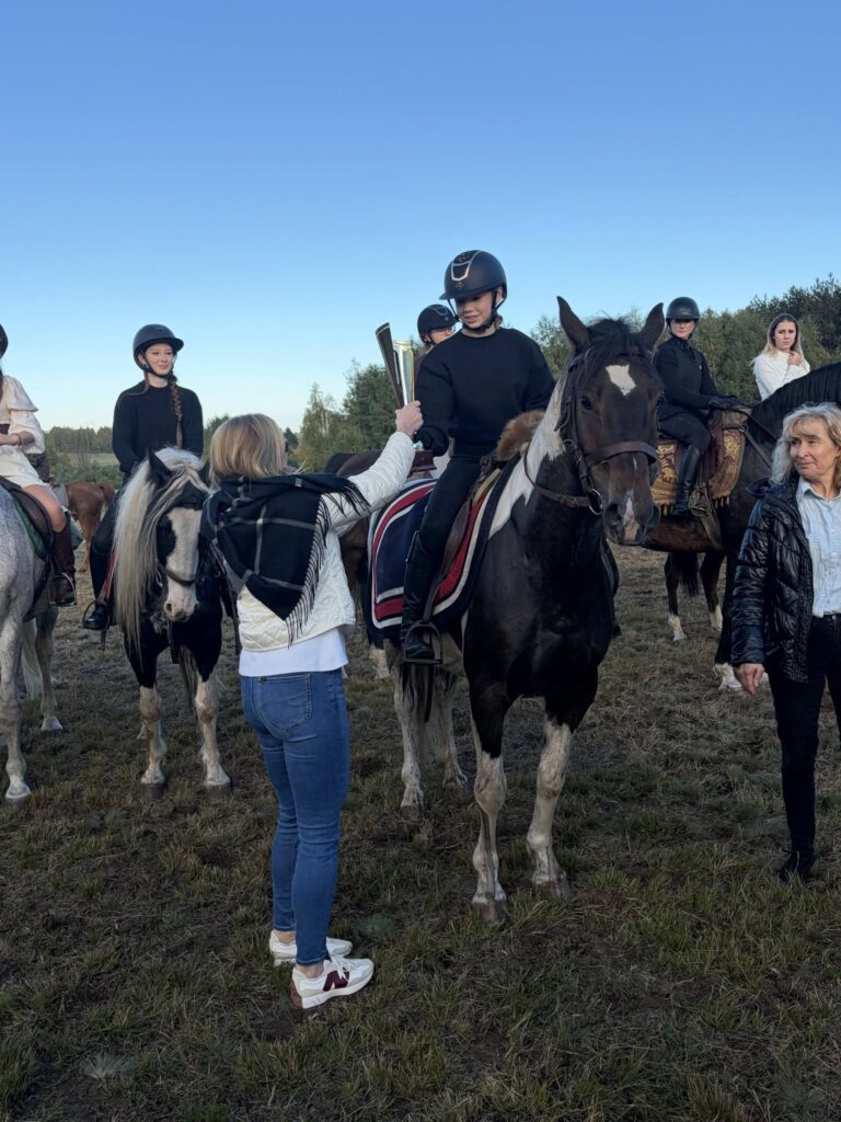 Na zdjęciu widzimy grupę młodych jeźdźców siedzących na koniach podczas uroczystego spotkania na świeżym powietrzu. W centrum fotografii znajduje się osoba w kasku i czarnym stroju, siedząca na dużym, ciemnym koniu z białymi znaczeniami na pysku i nogach. Na pierwszym planie stoi Pani skarbnik gminy Zakrzew ubrana w białą kurtkę, niebieskie dżinsy i sportowe buty. Wręcza jeźdźcowi puchar, za zwycięstwo w gonitwie za lisem podczas XIV Hubertusa w Gulinie. Wokół znajdują się inni uczestnicy na koniach o różnym umaszczeniu. Po prawej stronie stoi starsza kobieta w ciemnej kurtce, obserwująca przebieg wydarzenia.W tle widać drzewa, otwartą przestrzeń i błękitne, bezchmurne niebo. Konie mają założone siodła i ozdobne czapraki, a jeźdźcy noszą kaski ochronne.
