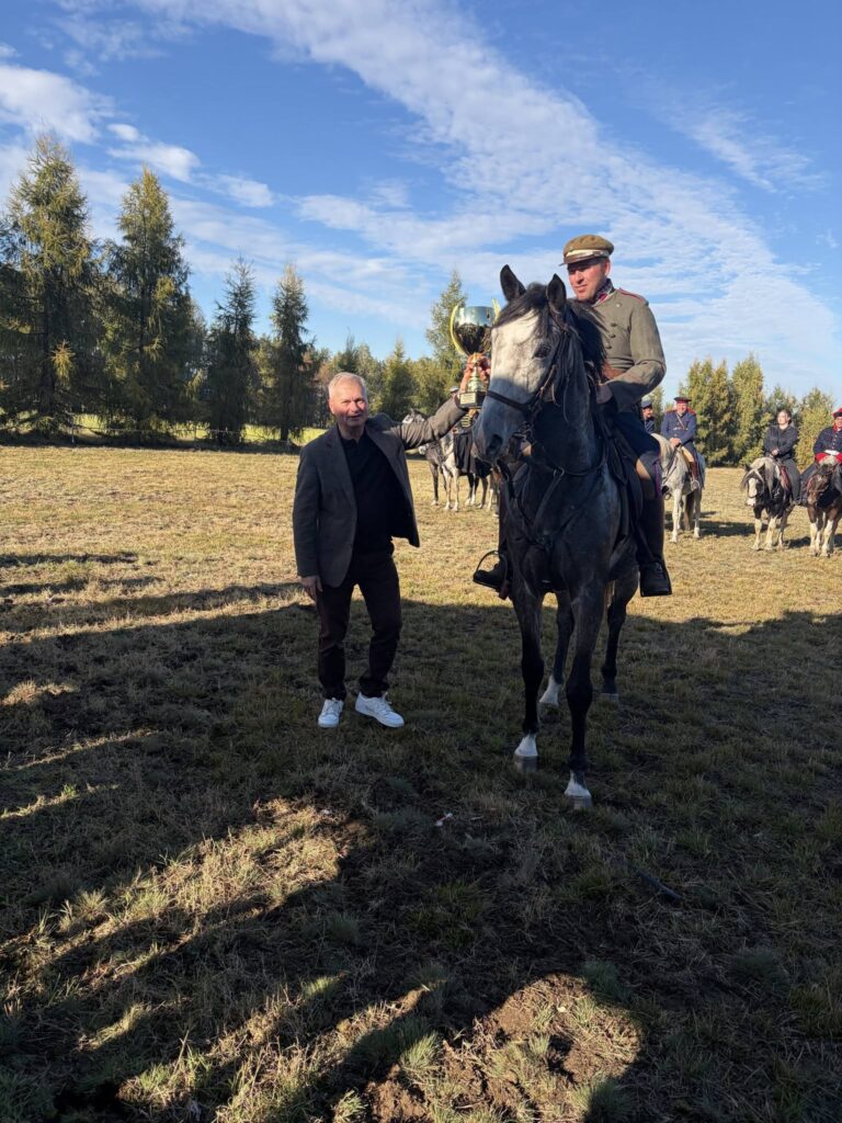 Na zdjęciu widzimy grupę młodych jeźdźców siedzących na koniach podczas uroczystego spotkania na świeżym powietrzu. W centrum fotografii znajduje się osoba, siedząca na dużym, ciemnym koniu. Na pierwszym planie stoi Pan wójt gminy Zakrzew ubrany w ciemną marynarkę i sportowe buty. Wręcza jeźdźcowi puchar, za zwycięstwo w gonitwie za lisem podczas XIV Hubertusa w Gulinie. W tle widać drzewa, otwartą przestrzeń i błękitne, bezchmurne niebo. Konie mają założone siodła i ozdobne czapraki, a jeźdźcy noszą kaski ochronne.