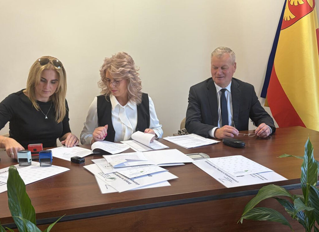 Trzy osoby siedzą przy stole podczas spotkania związanego z formalnościami dotyczącymi dofinansowania inwestycji wodociągowej w Gminie Zakrzew. Na stole znajdują się dokumenty, formularze i pieczątki. W tle widoczna jest flaga oraz elementy wyposażenia sali konferencyjnej.