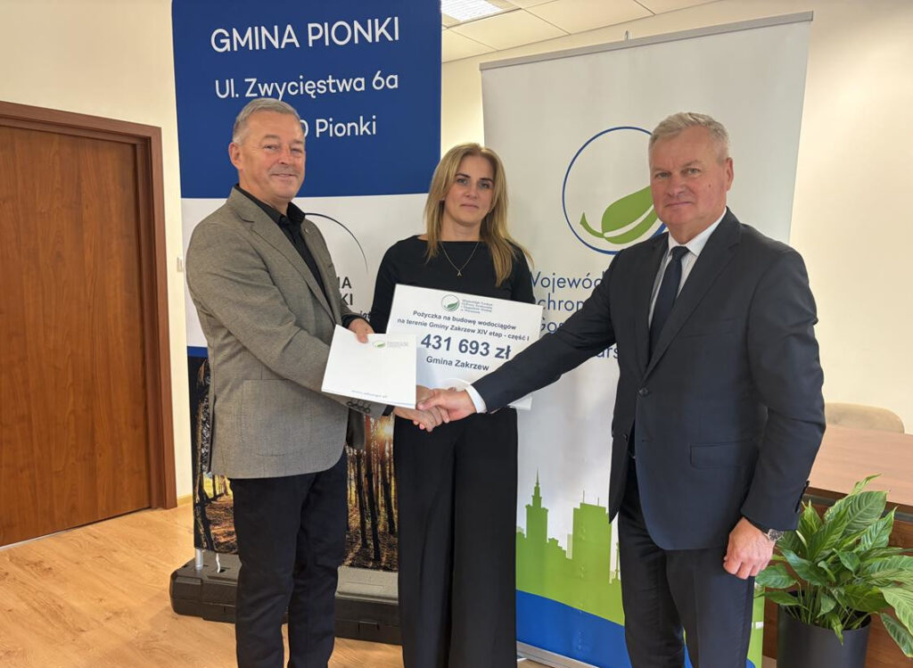 Trzy osoby prezentują dokument potwierdzający przyznanie dotacji w wysokości 431 693 zł na budowę wodociągu na terenie Gminy Zakrzew w ramach XIV etapu inwestycji. W tle widoczne są materiały promocyjne Gminy Pionki i Wojewódzkiego Funduszu Ochrony Środowiska i Gospodarki Wodnej.