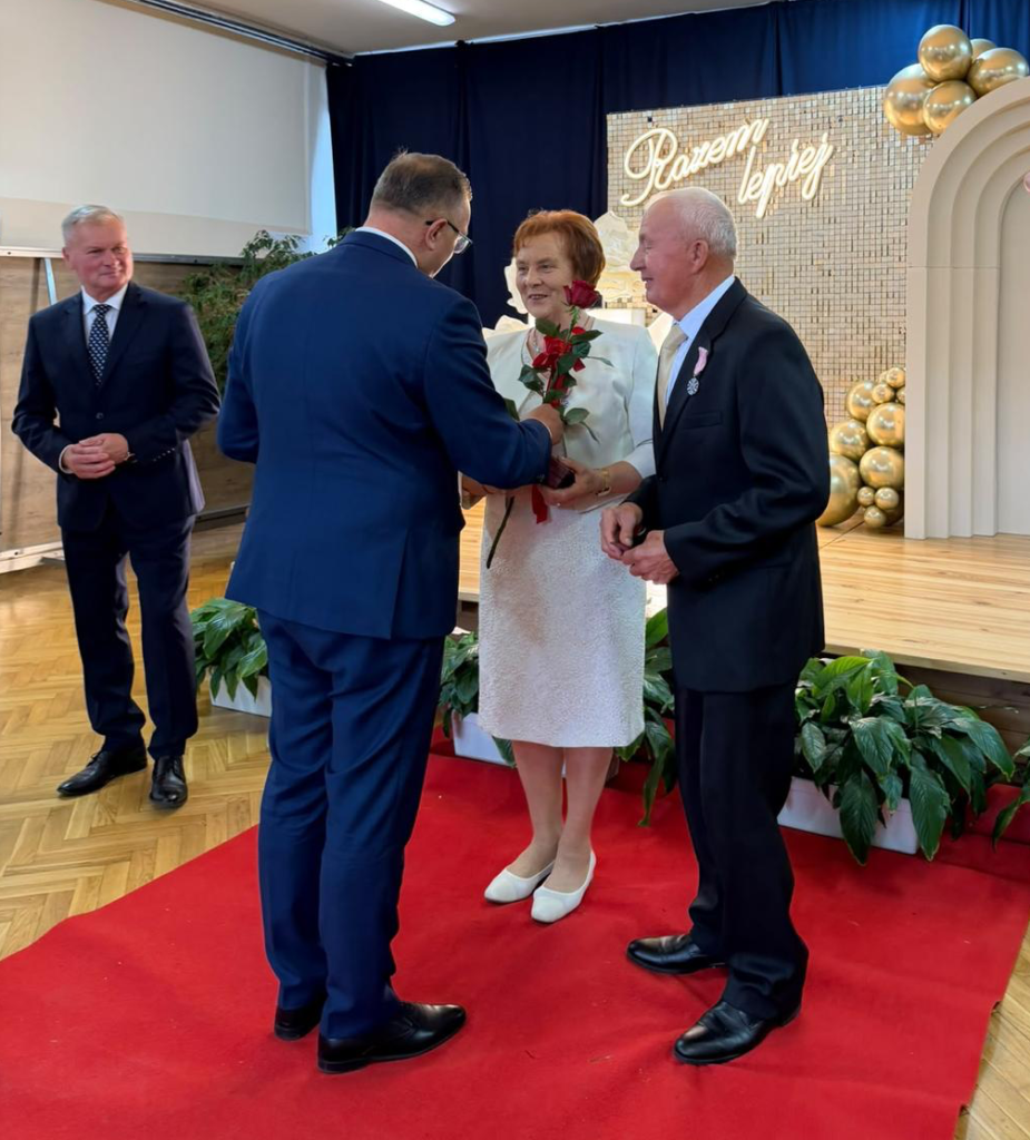 Podczas uroczystości jubileuszu długoletniego pożycia małżeńskiego przedstawiciel organizatorów wręcza jubilatce czerwoną różę. U jej boku stoi mąż, odznaczony medalem, z dumą i wzruszeniem uczestniczący w tej wyjątkowej chwili. W tle widoczny jest elegancki napis „Razem najlepiej”, który doskonale oddaje charakter spotkania.
To piękny gest uznania dla wspólnie przeżytych lat, wzajemnej miłości, wierności i przywiązania. Uroczystość przebiega w podniosłej, a zarazem serdecznej atmosferze, podkreślając, że prawdziwa miłość i trwałość małżeństwa zasługują na szczególne wyróżnienie.